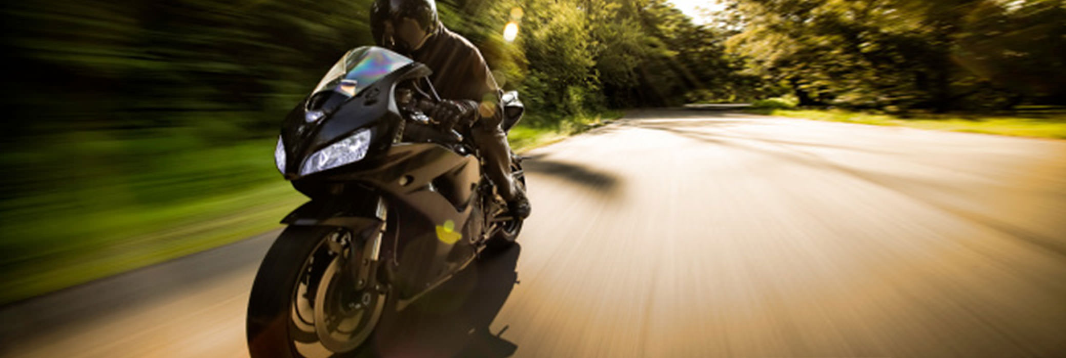 A guy in black riding his black motorcylce on a country road with trees at the sides